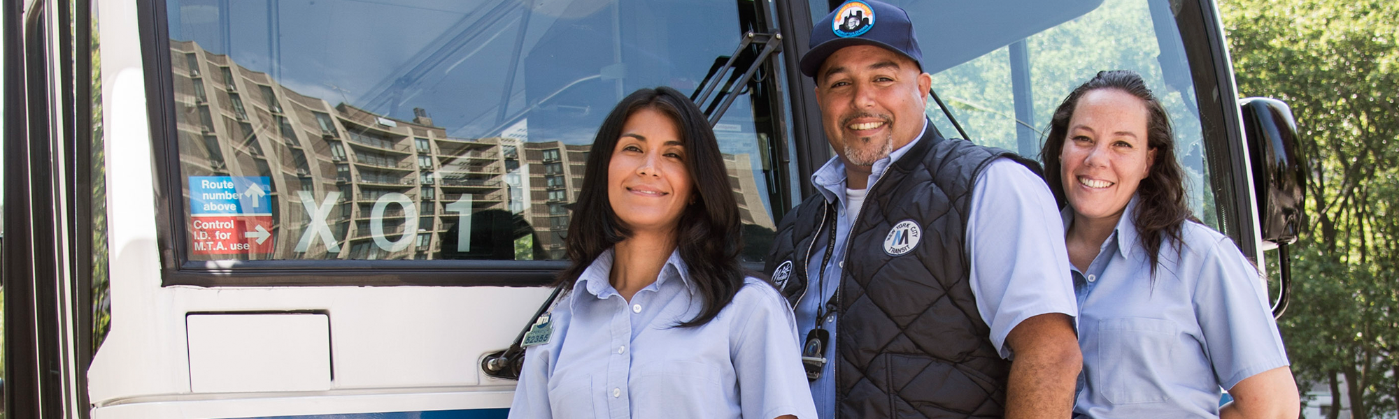 Three people stand in front of a bus. There are two women with brown hair wearing light blue shirts, and one man in a light blue shirt with a dark blue vest and a blue hat. 