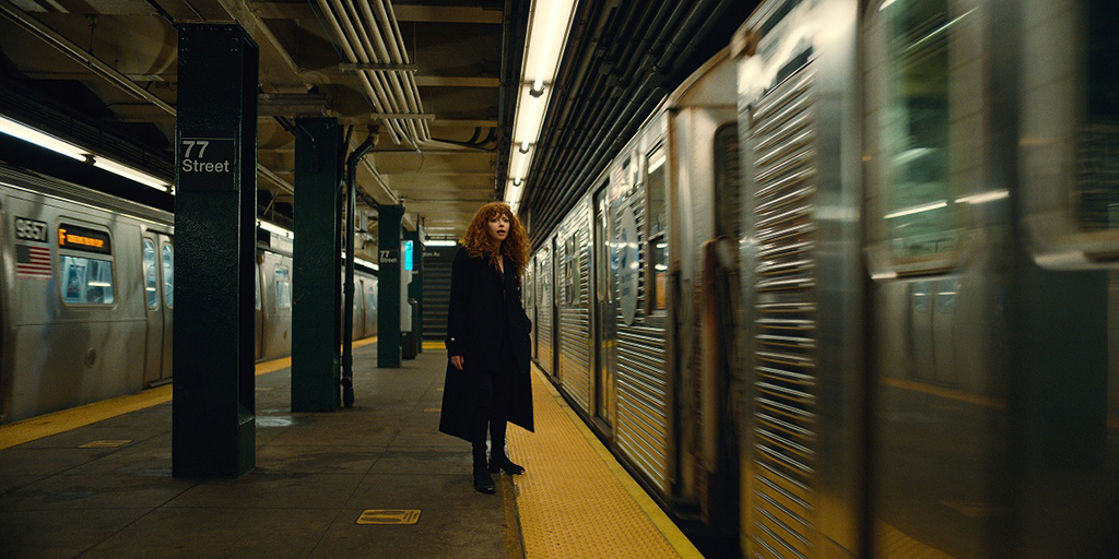 Natasha Lyonne, in a black coat, stands on a subway platform that says 77th St with two trains on either side