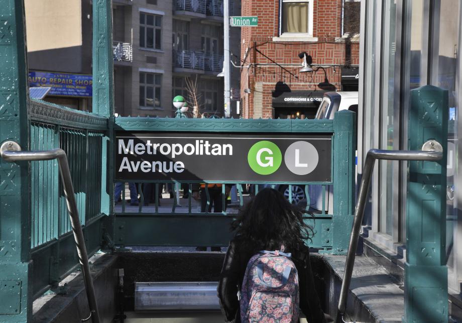 A woman with long, dark hair and a backpack walks down the stairs of a subway entrance. "Metropolitan Avenue" and the bullets for the G and the L trains are visible on a sign above the stairway.
