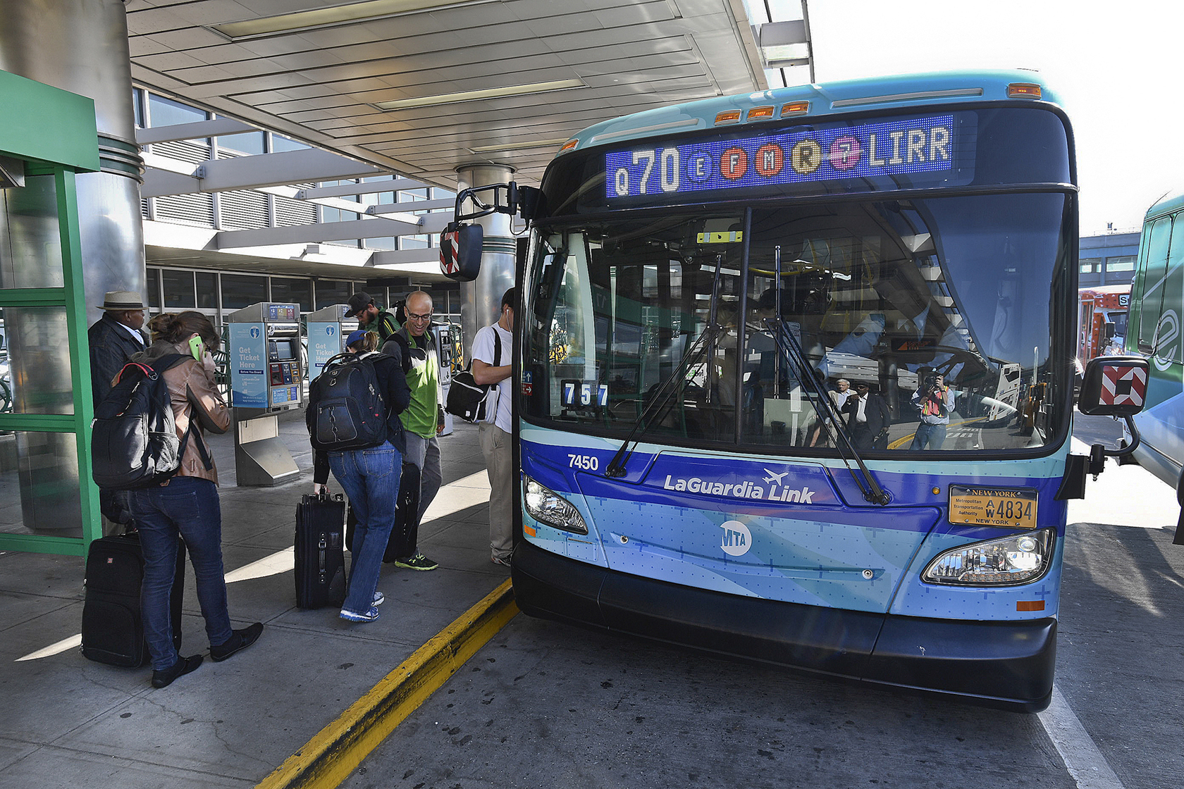 People with luggage wait to board a blue Q70 bus. The bus has a decal on the front that says LaGuardia Link.