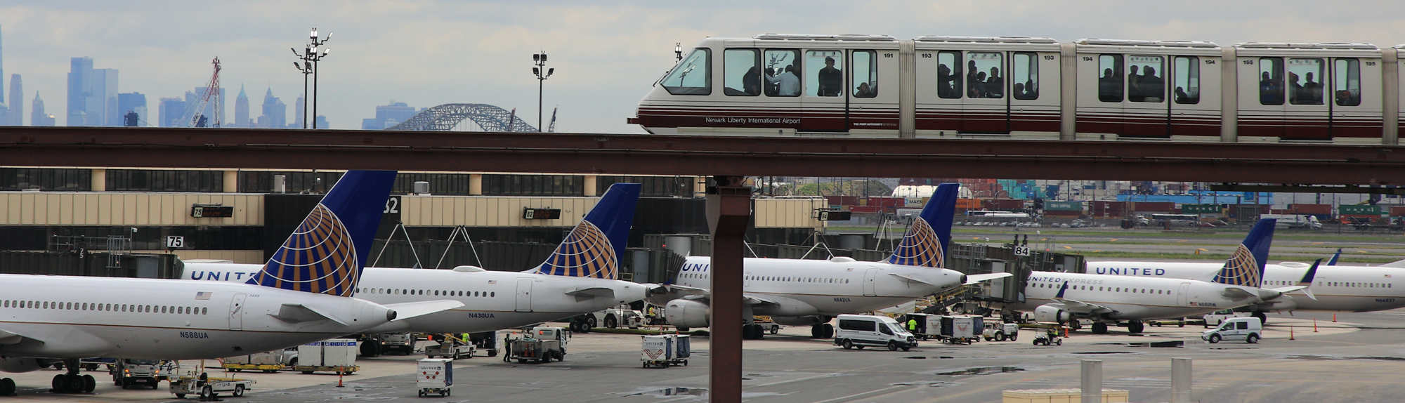 An AirTrain on an elevated track enters the frame on the right, with planes visible on the ground on the left. Newark Liberty International Airport is visible in the background.