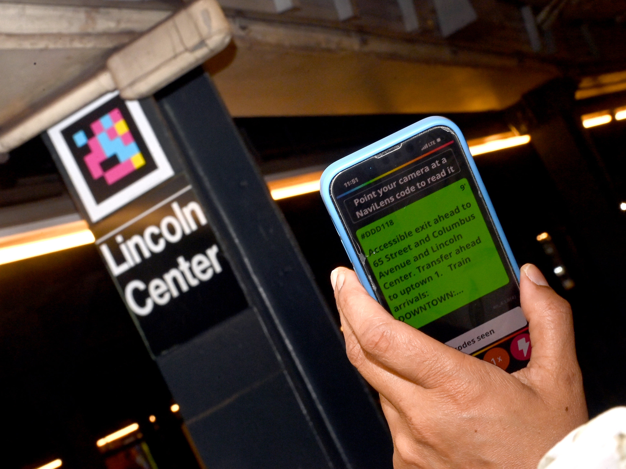 Phone in a hand pointed towards a NaviLens code in the background above a sign that says "Lincoln Center." The phone screen shows NaviLens information.