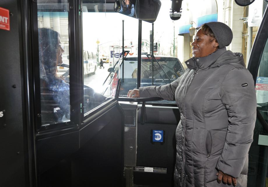 A woman wearing a coat and hat smiles at a bus operator as she inserts her MetroCard into the farebox at the front of a bus.