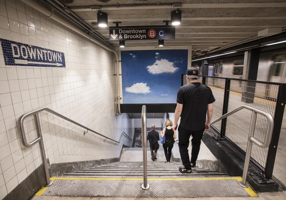 People walk down the stairs in a subway station. A sign reading "Downtown" is visible on the wall on the left, and a sign reading "Downtown & Brooklyn," with bullets for the B and C trains, is visible overhead.