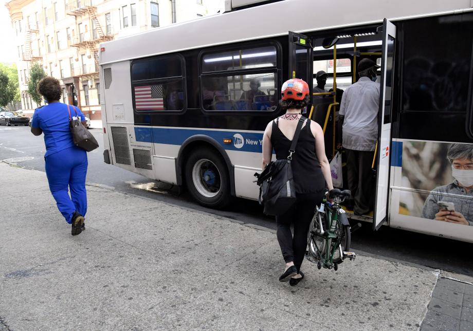 A woman carrying a green folding bike, currently folded up, approaches a bus at a bus stop.