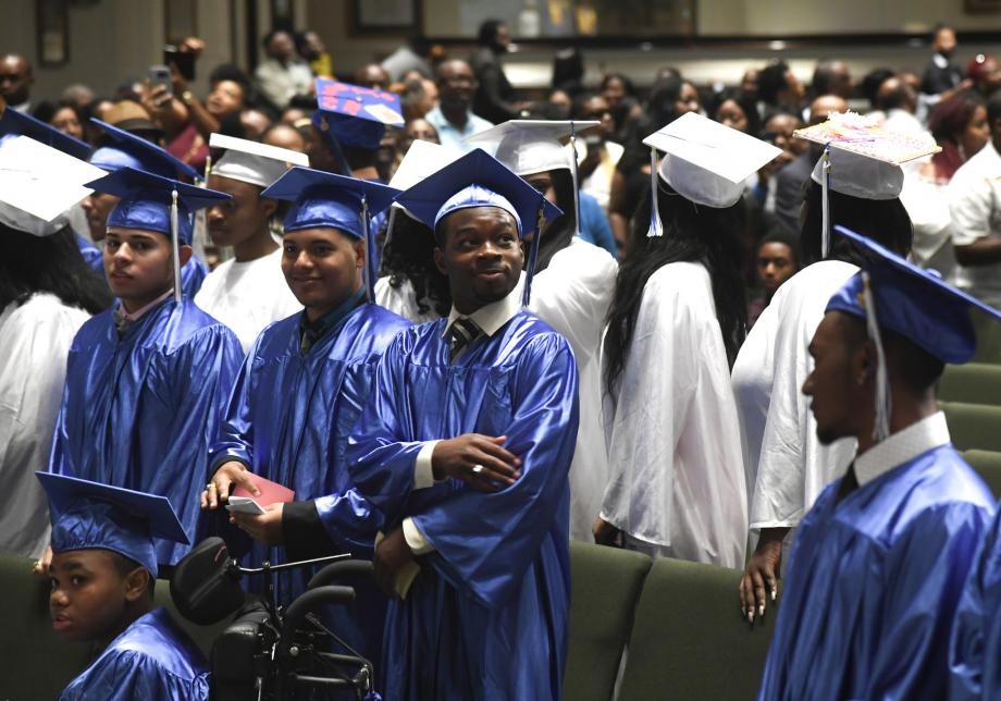 Students in blue and white graduation caps and gowns stand and sit in an auditorium. Many of them are smiling.