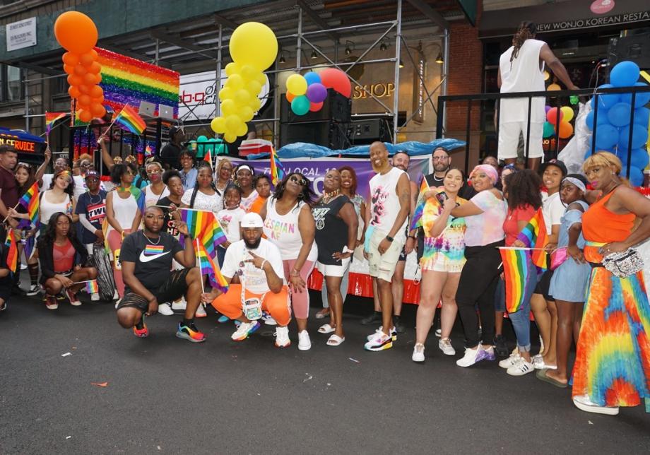 A big group of people, many wearing rainbow colors and carrying rainbow flags, smiles at the camera at a Pride Parade.