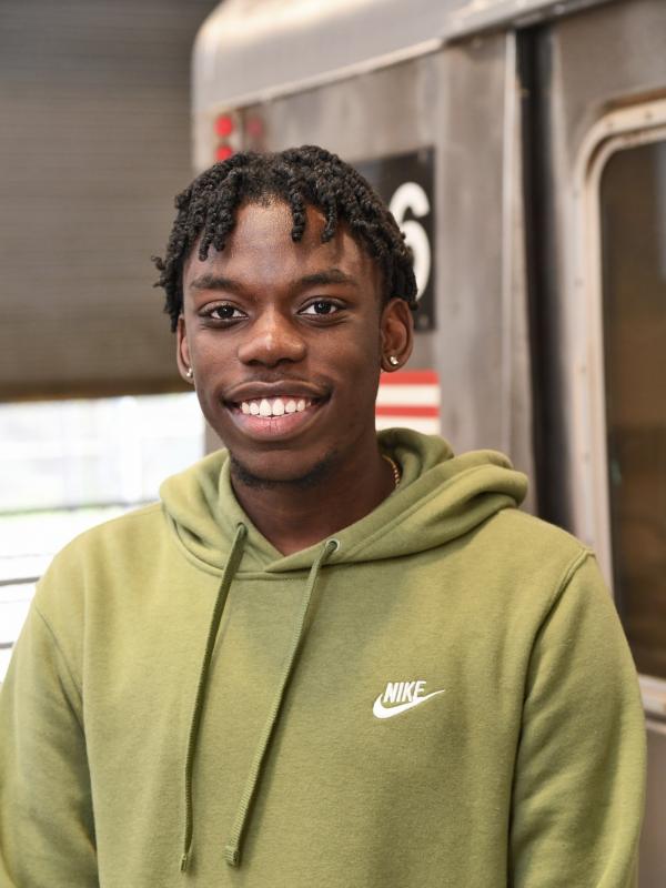 A young man with his hair in twists smiles at the camera, with a subway train car visible behind him.